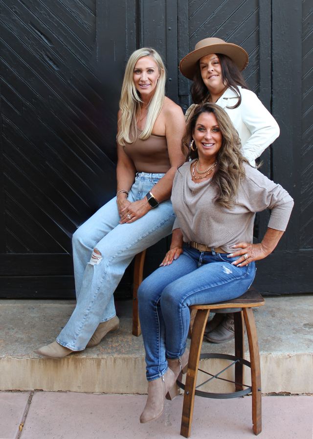 Barbie, Alison and Cynder of Suite 2 Salon posing together on wooden stools in front of a black wood door